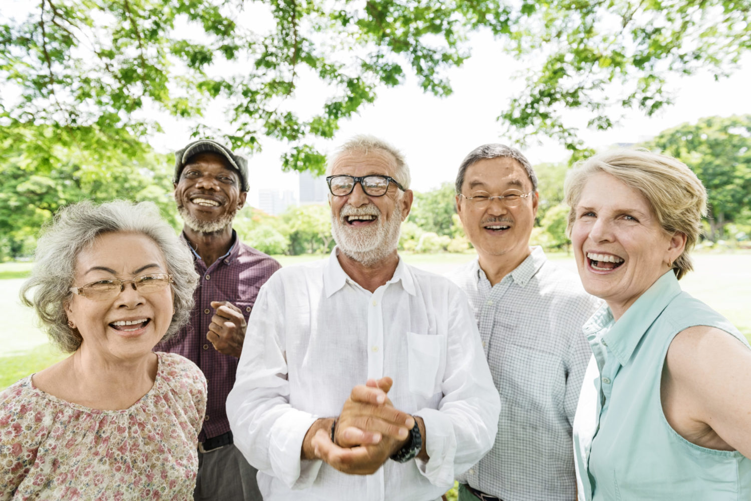 Tiefenbacher Group - A group of five older adults, three men and two women, stand close together outdoors, smiling and laughing under leafy trees on a sunny day, reflecting the joy that patient-centric drug development brings to thriving communities in a lush park setting.