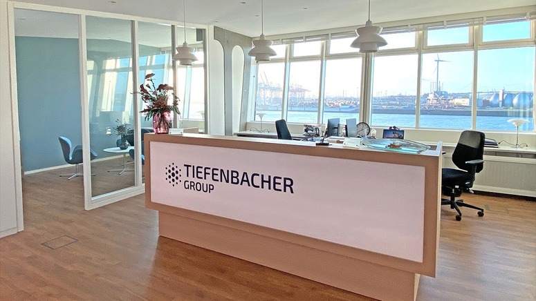 Tiefenbacher Group - A modern office reception area with a Tiefenbacher Group front desk, large windows overlooking an industrial port, wooden floors, and a glass-walled meeting room—reflecting their focus on patient-centric drug development.