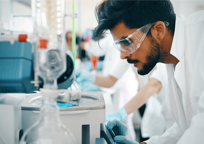 Tiefenbacher Group - A scientist wearing safety goggles and a lab coat closely observes equipment in a laboratory focused on patient-centric drug development, surrounded by scientific instruments and glassware.