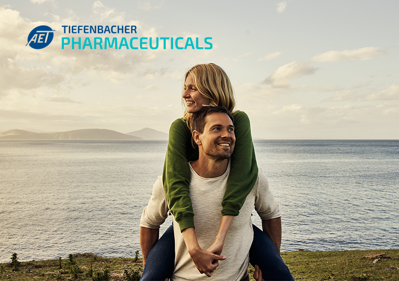 Tiefenbacher Group - A smiling man gives a piggyback ride to a woman by the sea, both looking happy. The TIEFENBACHER PHARMACEUTICALS logo appears in the top left corner, representing family-owned pharmaceutical company values. The background shows water, grass, and a cloudy sky.