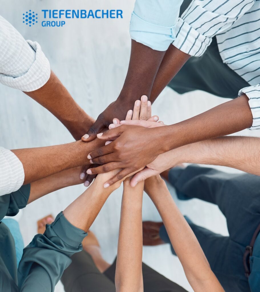 Tiefenbacher Group - A group of diverse people stand in a circle, stacking their hands to symbolize unity and teamwork. The TIEFENBACHER GROUP logo appears in the top left, reflecting their role as a family-owned pharmaceutical company specializing in finished dosage forms.