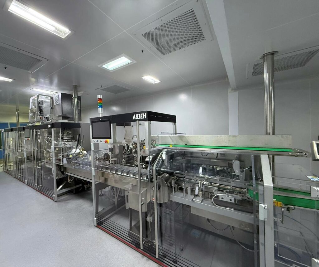 Tiefenbacher Group - A modern packaging machine for pharmaceutical healthcare solutions operates in a clean, well-lit factory with gray floors, white ceilings, transparent panels, conveyor belts, and colored indicator lights.