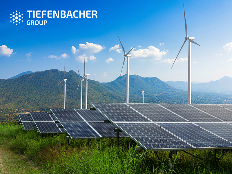 Tiefenbacher Group - Solar panels and wind turbines in a green field with mountains in the background under a blue sky, highlighting TIEFENBACHER GROUP’s commitment to patient-centric drug development. The TIEFENBACHER GROUP logo appears in the top left corner.