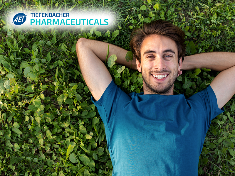 Tiefenbacher Group - A smiling young man in a blue shirt lies on his back in green grass with his hands behind his head, reflecting the patient-centric drug development focus of Tiefenbacher Pharmaceuticals. The company’s logo appears in the top left corner.