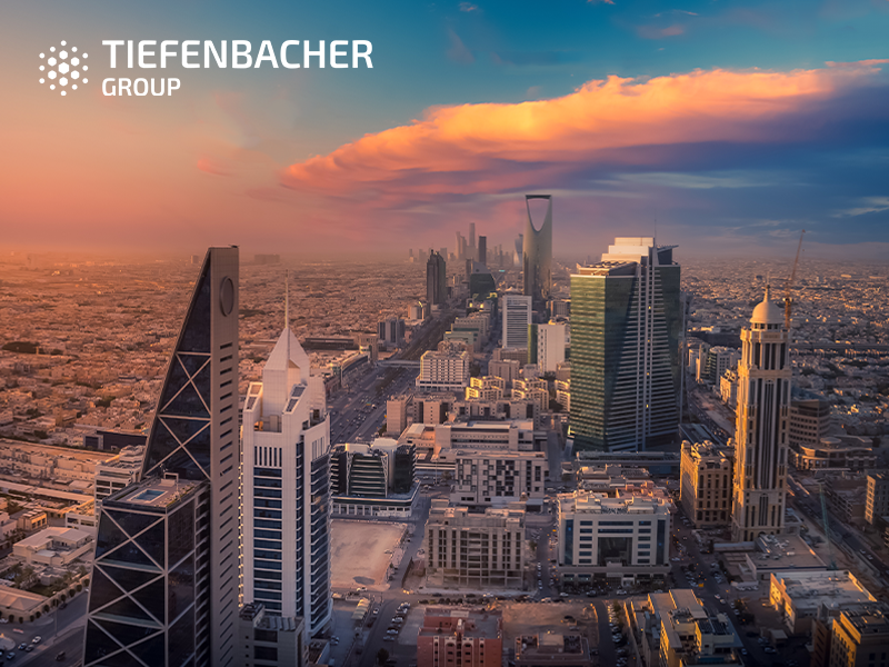 Tiefenbacher Group - Aerial view of Riyadh city skyline at sunset with modern skyscrapers, including the Kingdom Centre, under a dramatic sky. "Tiefenbacher Group" logo is at the top left, representing pharmaceutical healthcare solutions.