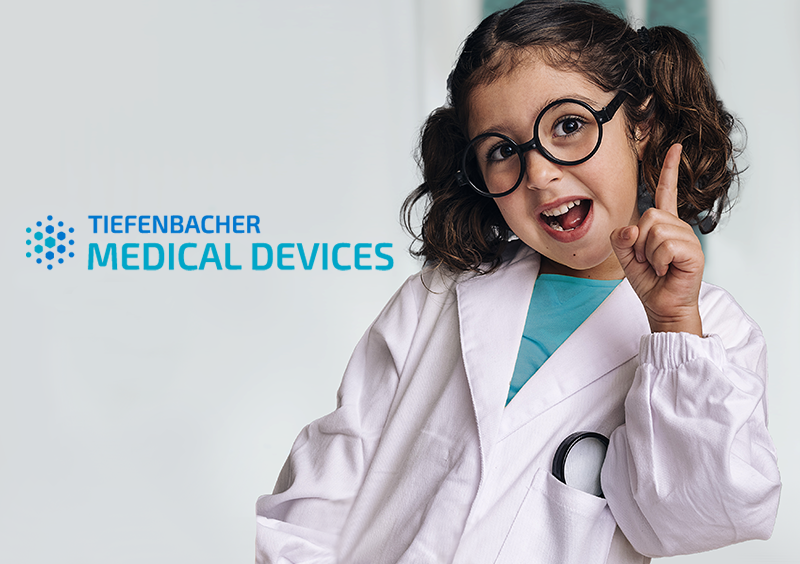 Tiefenbacher Group - A young girl wearing glasses and a white lab coat smiles and points upward, standing next to the text "Tiefenbacher Medical Devices," representing innovative pharmaceutical healthcare solutions on a light background.