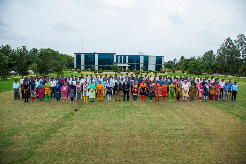 Tiefenbacher Group - A large group of people, including men and women in formal and colorful traditional attire, stand on a grassy lawn with a modern glass building behind them, reflecting the unity of a family-owned pharmaceutical company dedicated to patient-centric drug development.