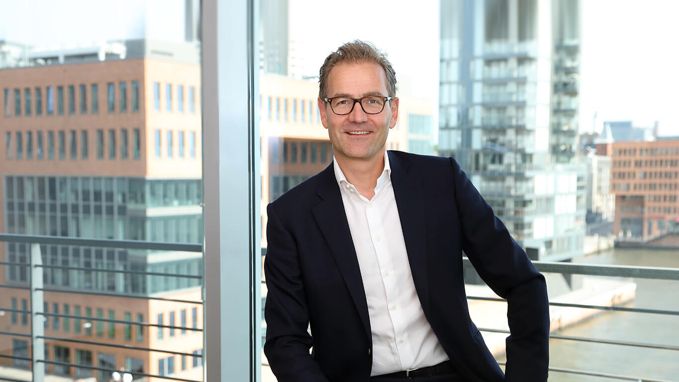 Tiefenbacher Group - A man in a dark suit and glasses sits on a chair by a large window overlooking modern office buildings and a waterfront. He is smiling, has short, light hair, and projects the professionalism of a family-owned pharmaceutical company.