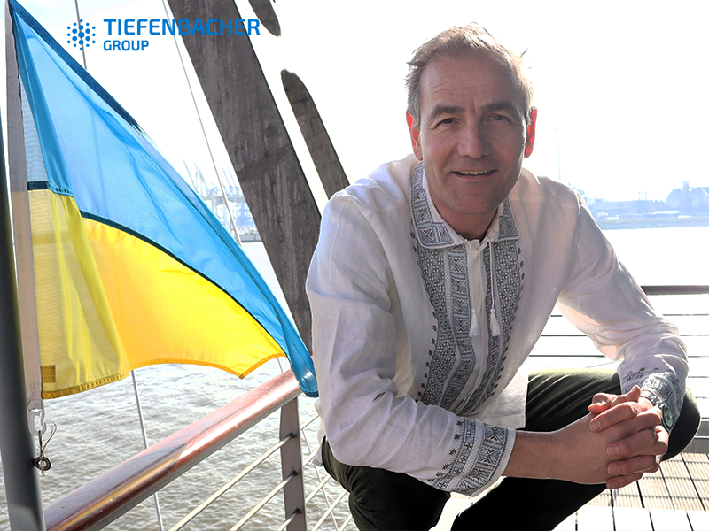 Tiefenbacher Group - A man in a traditional embroidered shirt kneels on a riverside deck next to a Ukrainian flag. The Tiefenbacher Group logo, a symbol of patient-centric drug development and pharmaceutical healthcare solutions, is in the upper left corner. Sunlight shines behind him.