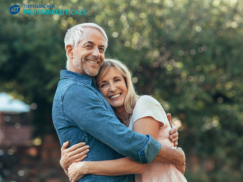 Tiefenbacher Group - A smiling older couple embraces outdoors in a garden, looking happy and relaxed. The man wears a blue shirt; the woman, a light pink top. The "Tiefenbacher Pharmaceuticals" logo highlights their family-owned pharmaceutical company in the upper left corner.