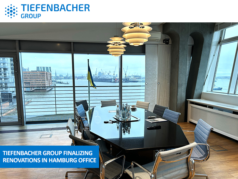 Tiefenbacher Group - Modern office conference room with a round table, chairs, and large windows overlooking a river and cityscape. Overhead lights illuminate the space. A notification reads, "Family-owned pharmaceutical company Tiefenbacher Group finalizing renovations in Hamburg office.