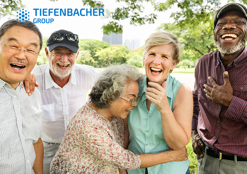 Tiefenbacher Group - A diverse group of five older adults stand outdoors in a park, laughing and smiling together. The Tiefenbacher Group logo is displayed in the top left corner, reflecting their commitment to innovative pharmaceutical healthcare solutions.