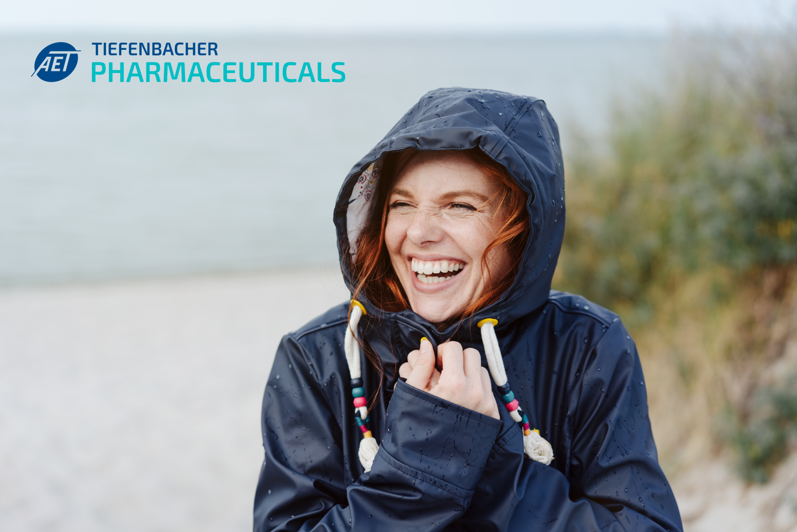 Tiefenbacher Group - A smiling woman in a navy raincoat stands on a beach, holding her hood. The AET TIEFENBACHER PHARMACEUTICALS logo appears in the top left, reflecting their focus on patient-centric drug development. The background shows the sea and greenery.
