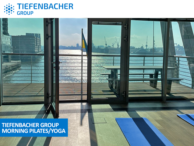 Tiefenbacher Group - Large windows overlook a river and cityscape with cranes in the distance. Inside, blue yoga mats are laid out on the floor, sunlight streaming in, with a sign reading "Tiefenbacher Group Morning Pilates/Yoga," reflecting their commitment to pharmaceutical healthcare solutions.