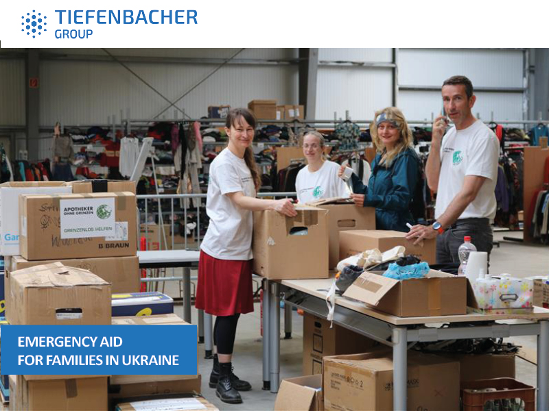 Tiefenbacher Group - Four volunteers in a warehouse sort and pack boxes of donated goods for families in Ukraine. Shelves and tables are filled with supplies, and the Tiefenbacher Group logo highlights their commitment to pharmaceutical healthcare solutions.