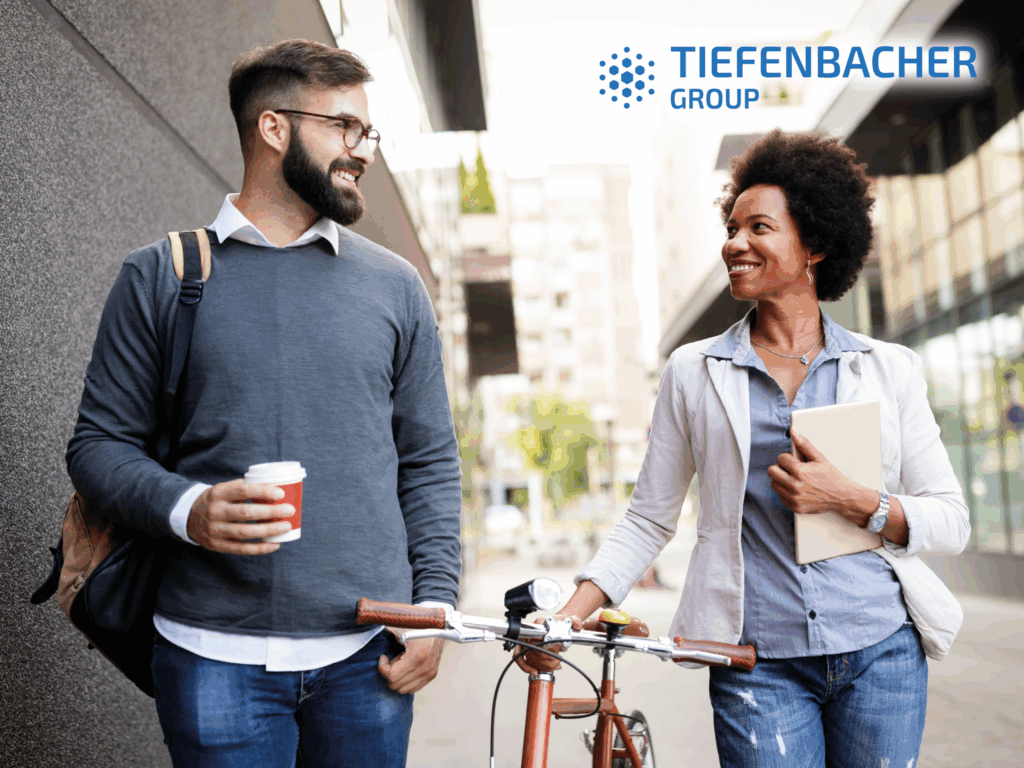 Tiefenbacher Group - A man holding a coffee cup and a woman with a tablet walk together outdoors, smiling at each other, as she walks a bicycle. The TIEFENBACHER GROUP logo in the top right highlights this family-owned pharmaceutical company specializing in finished dosage forms.