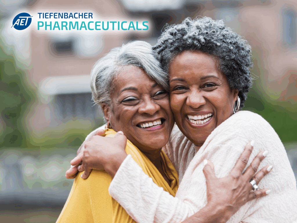 Tiefenbacher Group - Two older women smiling and hugging outdoors, both appearing joyful. The logo and text “AET TIEFENBACHER PHARMACEUTICALS” is visible in the top left corner, highlighting patient-centric drug development.