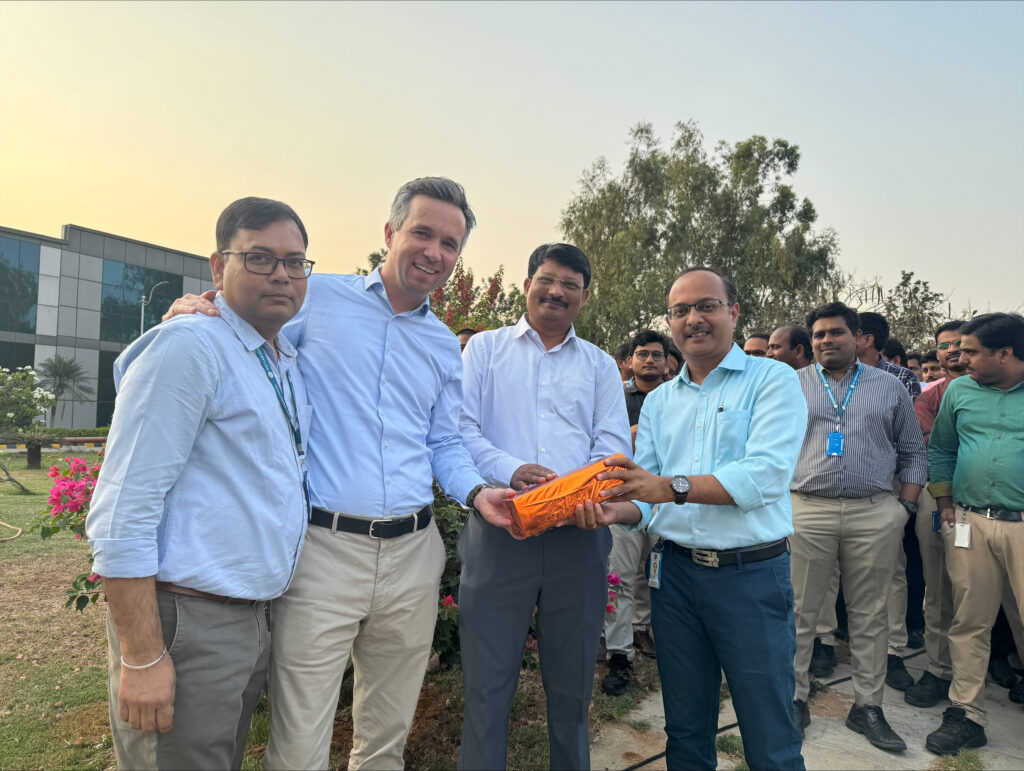 Tiefenbacher Group - A group of men, some holding an orange rope, stand outdoors in front of blooming flowers and a modern building, smiling for a photo as colleagues from a pharmaceutical healthcare solutions team gather behind them.