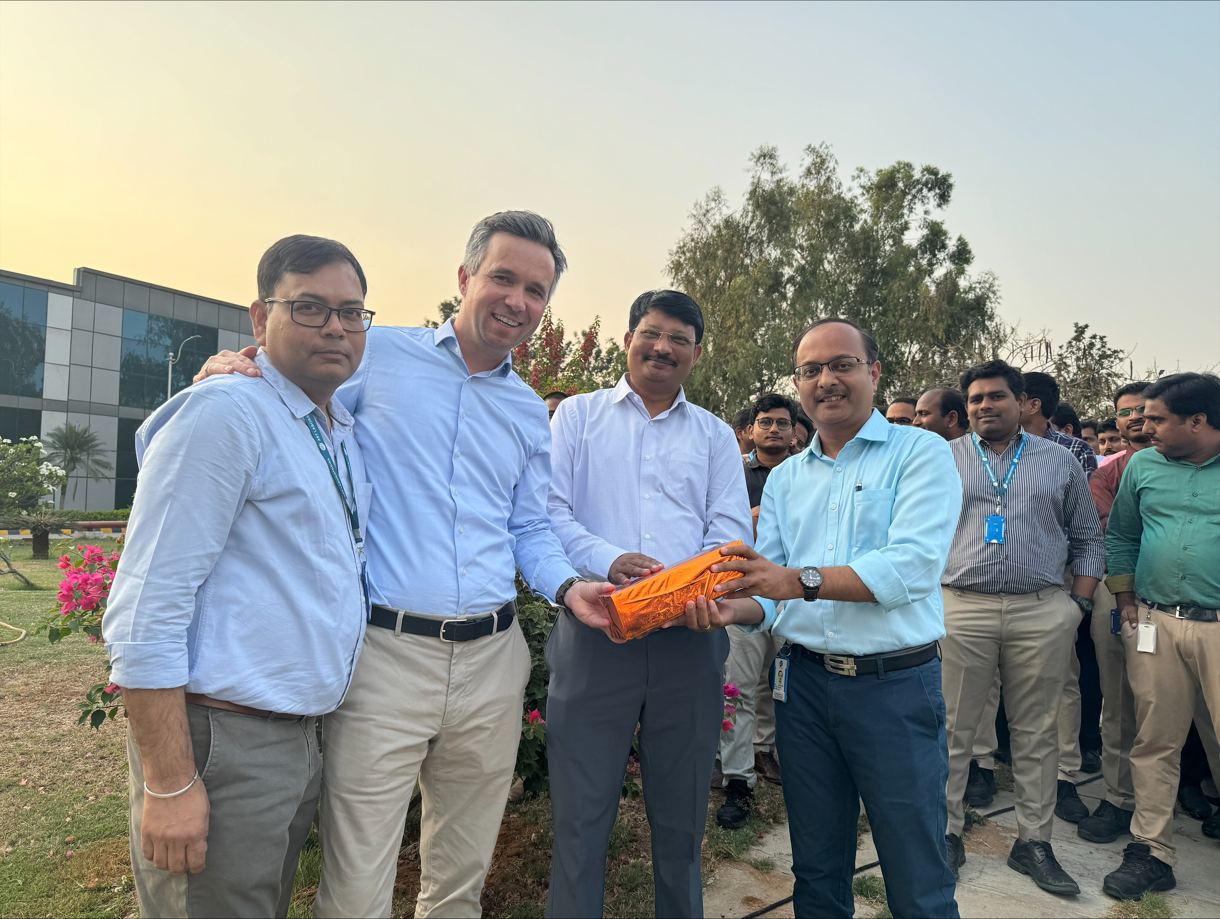 Tiefenbacher Group - A group of men, some holding an orange rope, stand outdoors in front of blooming flowers and a modern building, smiling for a photo as colleagues from a pharmaceutical healthcare solutions team gather behind them.