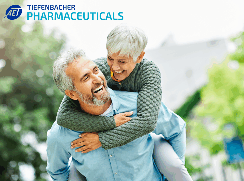 Tiefenbacher Group - A smiling older woman with short gray hair joyfully gives a piggyback ride to a laughing older man outdoors, highlighting Tiefenbacher Pharmaceuticals' commitment to generic medicines manufacturing. The company logo is in the top left corner.